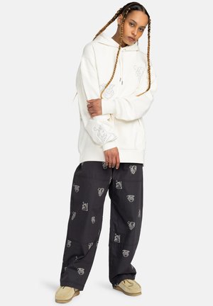 Oversized cream hoodie with graphic print, paired with dark trousers featuring repeated white patterns. Model wears beige slip-on shoes.