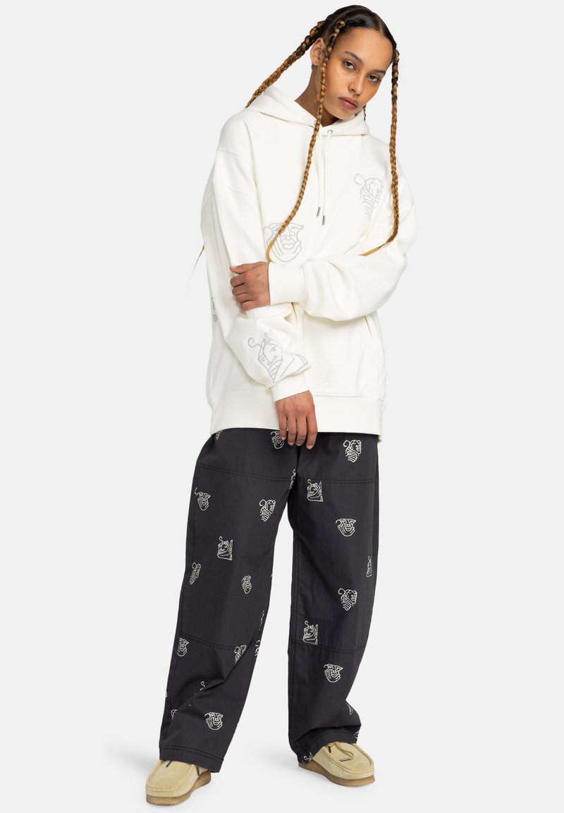 Oversized cream hoodie with graphic print, paired with dark trousers featuring repeated white patterns. Model wears beige slip-on shoes.
