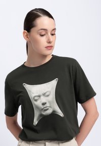 Donkergroene katoenen t-shirt met een opvallende witte gezichtsafbeelding die is ingekapseld in een textuurpatch met contouren en zilveren hardwareaccenten.