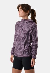 Žena smeđom kosom zavezanoj u rep, odjevena u ljubičastu maskirnu jaknu na patentni zatvarač i crne šortse, okrenuta lijevo na bijeloj pozadini.