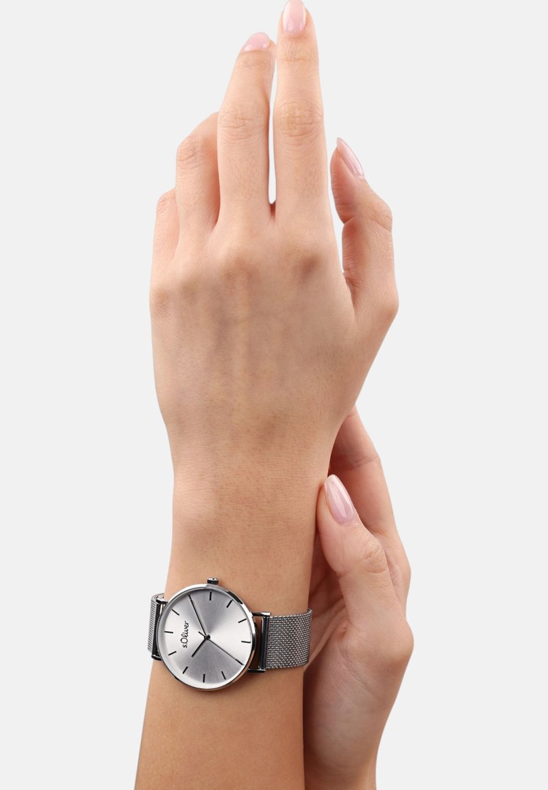 Zilveren ronde polshorloge met een mesh band. Het heeft een gladde, reflecterende wijzerplaat met minimalistische uurmarkeringen en branding.