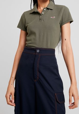 Vrouw draagt een olijfgroen poloshirt met korte mouwen en marineblauwe broek met hoge taille, oranje stiksels en cargozak.
