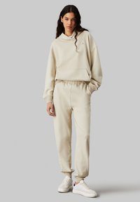 Sudadera beige de corte relajado con capucha y bolsillo delantero, combinada con joggers a juego que tienen puños elásticos. Zapatillas blancas con detalles.