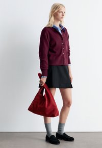 Burgunderfarbener Cardigan mit Knöpfen, hellblaues Hemd darunter, schwarzer Rock, graue Socken, schwarze Loafers und eine rote Wildleder-Schultertasche.