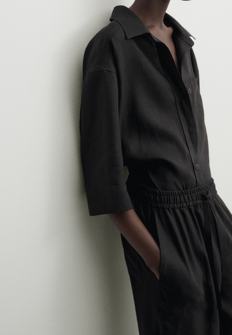 Personne portant une chemise noire ample à boutons avec col et un pantalon noir à taille élastique, debout avec une main dans la poche contre un mur clair.