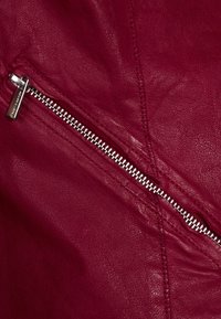 Rote Lederjacke mit silbernem Reißverschlussdetail. Die Textur ist glatt mit sichtbaren Nähten; der Reißverschluss verläuft diagonal über den Stoff.