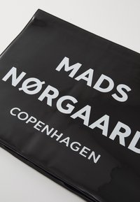 Schwarze, glänzende Tragetasche mit auffälligem weißen Text, der "MADS NØRGAARD COPENHAGEN" lautet. Glatte Textur ohne weitere Muster oder Akzente.