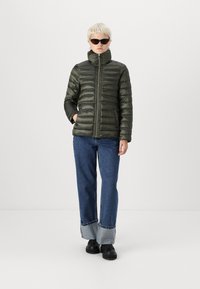 Olivengrønn pufferjakke med vertikal quiltet mønster, høy krage, frontlås, parret med løse blå jeans og svarte sko.