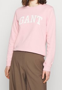 Sweat-shirt rose clair en mélange de coton doux. Il présente le lettrage blanc "GANT" sur la poitrine et des poignets côtelés avec une coupe décontractée.