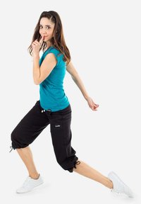 Pantaloni neri a vita alta con polsini regolabili, abbinati a una maglietta aderente turchese. Il modello è in una posa dinamica, indossando sneaker bianche.