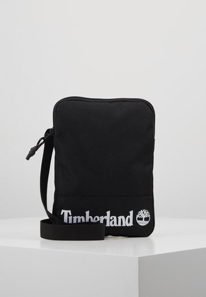 Zwarte rechthoekige crossbodytas met verstelbare riem, ritssluiting en wit Timberland-logo en boomembleem aan de voorzijde onderaan.