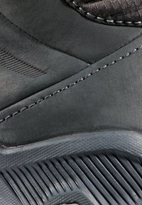 Nahaufnahme eines schwarzen Sneakers mit strukturierter Gummisohle, Wildleder-Obermaterial mit Nahtdetails und gepolstertem Kragen mit Stoffstruktur.