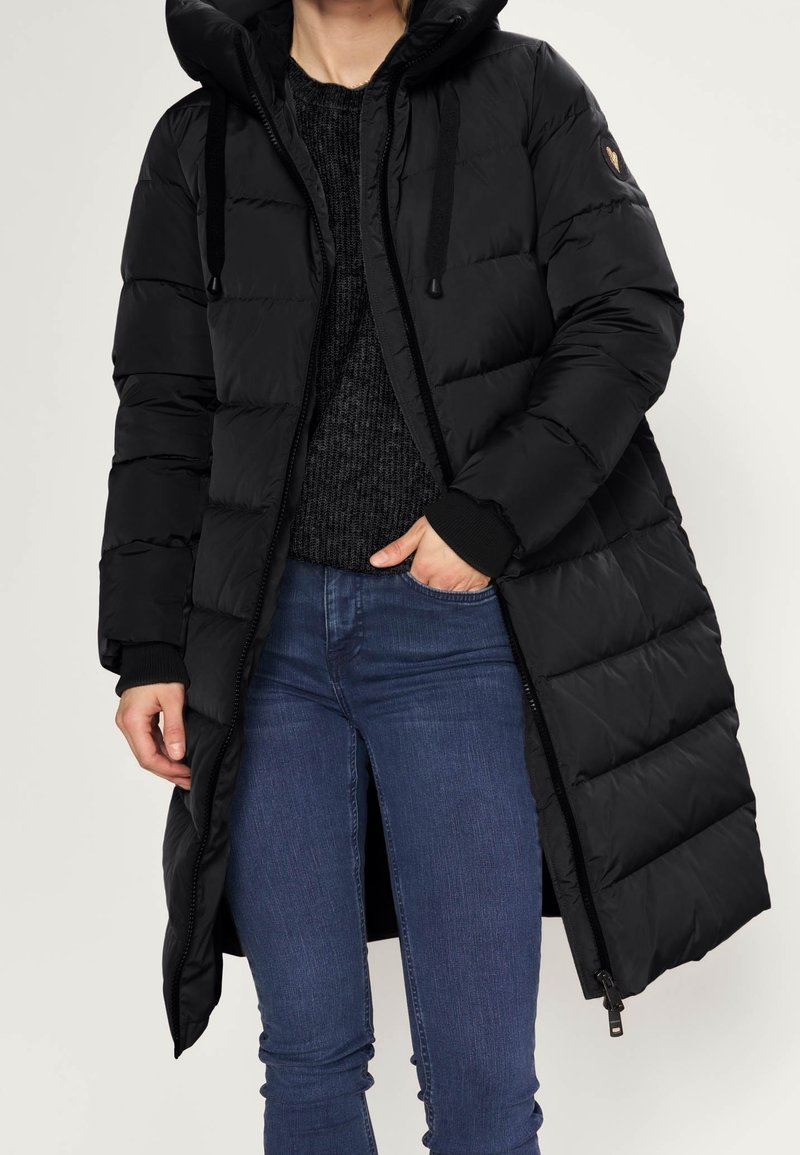 Person som bär en lång svart vadderad kappa med huva, svart stickad tröja och blå jeans, handen i fickan, mot en enfärgad bakgrund.