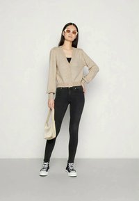 Mulher vestindo um cardigan bege com botões, jeans skinny pretos, ténis altos pretos, óculos de sol brancos, segurando uma bolsa beje, em frente a um fundo branco.