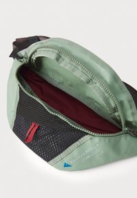 Belt bag v zeleném materiálu s síťovými akcenty, kontrastními černými částmi, červeným táhlem na zip a vínově červenou podšívkou. Otevřený k nahlédnutí.