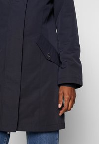 Marinblå vattentät jacka med upprätt krage, framficka med knäppning och justerbara ärmslut. Slät textur och slitstark tyg.