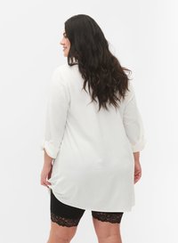 Nainen, jolla on pitkät, aaltoilevat tummat hiukset, valkoinen pitkähihainen tunika ja mustat pitsireunaiset pyöräilijäshortsit, seisoo valkoista taustaa vasten.