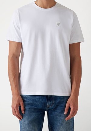 T-shirt en coton blanc avec un col rond et des manches courtes, orné d'un petit logo triangulaire sur la poitrine. Porté avec un jean bleu.