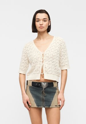 Jonge vrouw die een crèmekleurige, gebreide, korte cardigan met linten draagt en een tweekleurige denim minirok met een gesp in de vorm van een hart.