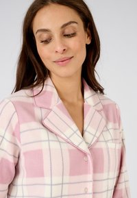 Camisa de pijama de franela de cuadros en rosa y blanco con cuello, cierre de botones y bolsillo en el pecho, que presenta una textura suave y líneas grises contrastantes.