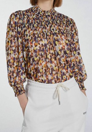 Femme portant un chemisier à manches longues fleuri coloré avec un col smocké et des poignets, associé à un pantalon blanc à cordon avec poches.