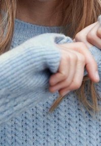 Maglione azzurro chiaro a maglia con un motivo testurizzato e fori per le dita, mentre le mani regolano le maniche. Materiale morbido e accogliente.