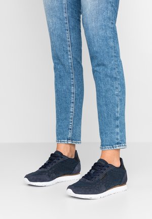 Person wearing blue slim-fit jeans and dark blue lace-up sneakers with white soles, standing on a plain light gray floor.