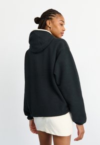 Μαύρη φλις κουκούλα με υφή, υπερμεγέθους στυλ και ρυθμιζόμενη κουκούλα. Φορεμένη πάνω από ένα ανοιχτό κρεμ πουκάμισο.