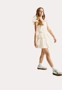 Vit, knälång klänning med volangärmar och lagerdesign. Matchad med silverfärgade sneakers och vita strumpor. Slät tygtextur.