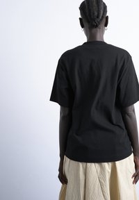 Personne avec un chignon tressé portant des boucles d'oreilles en argent en forme de créole, un t-shirt noir à manches courtes et une jupe plissée beige, vue de dos sur fond blanc.