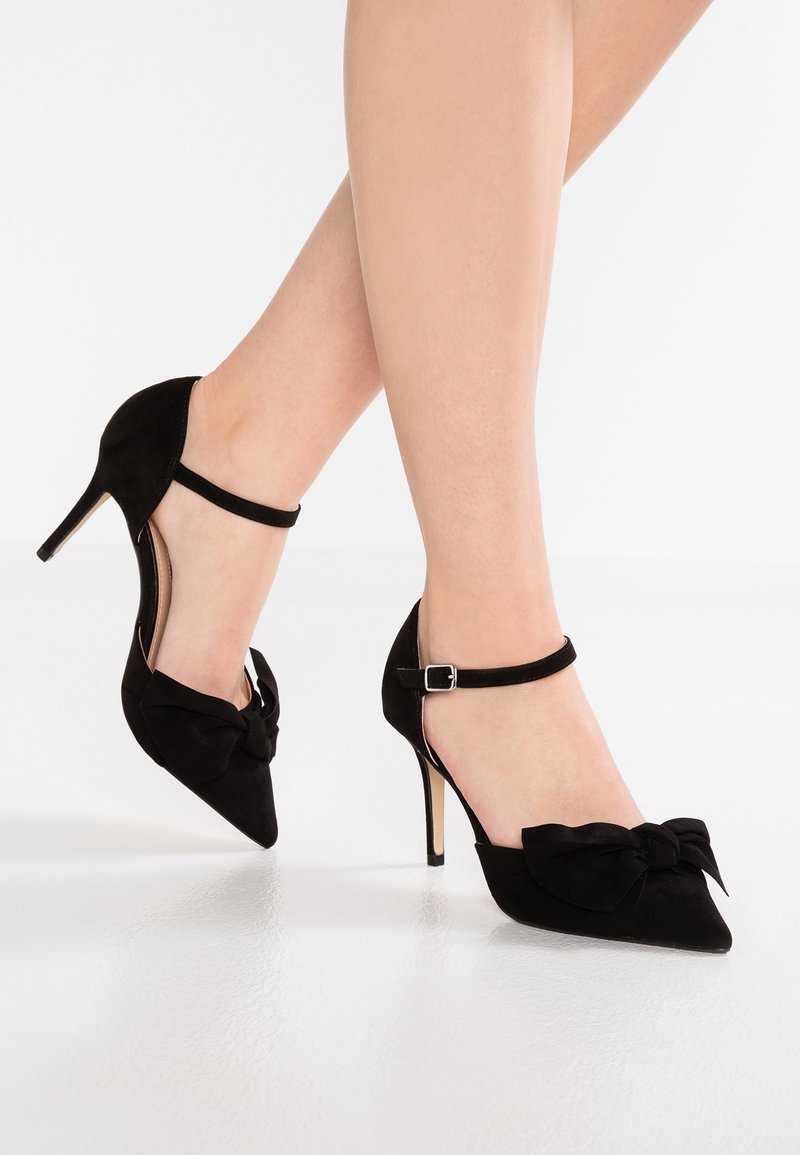 Femme portant des talons aiguilles noirs à bouts pointus avec des brides à la cheville et de grands nœuds décoratifs, debout sur un sol blanc.