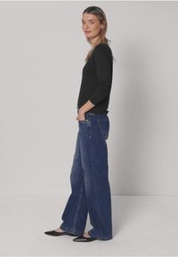 Maglia a maniche lunghe scura, abbinata a jeans blu a vita alta e gamba larga. Ballerine nere con punta a punta. Modella in piedi di profilo su uno sfondo neutro.