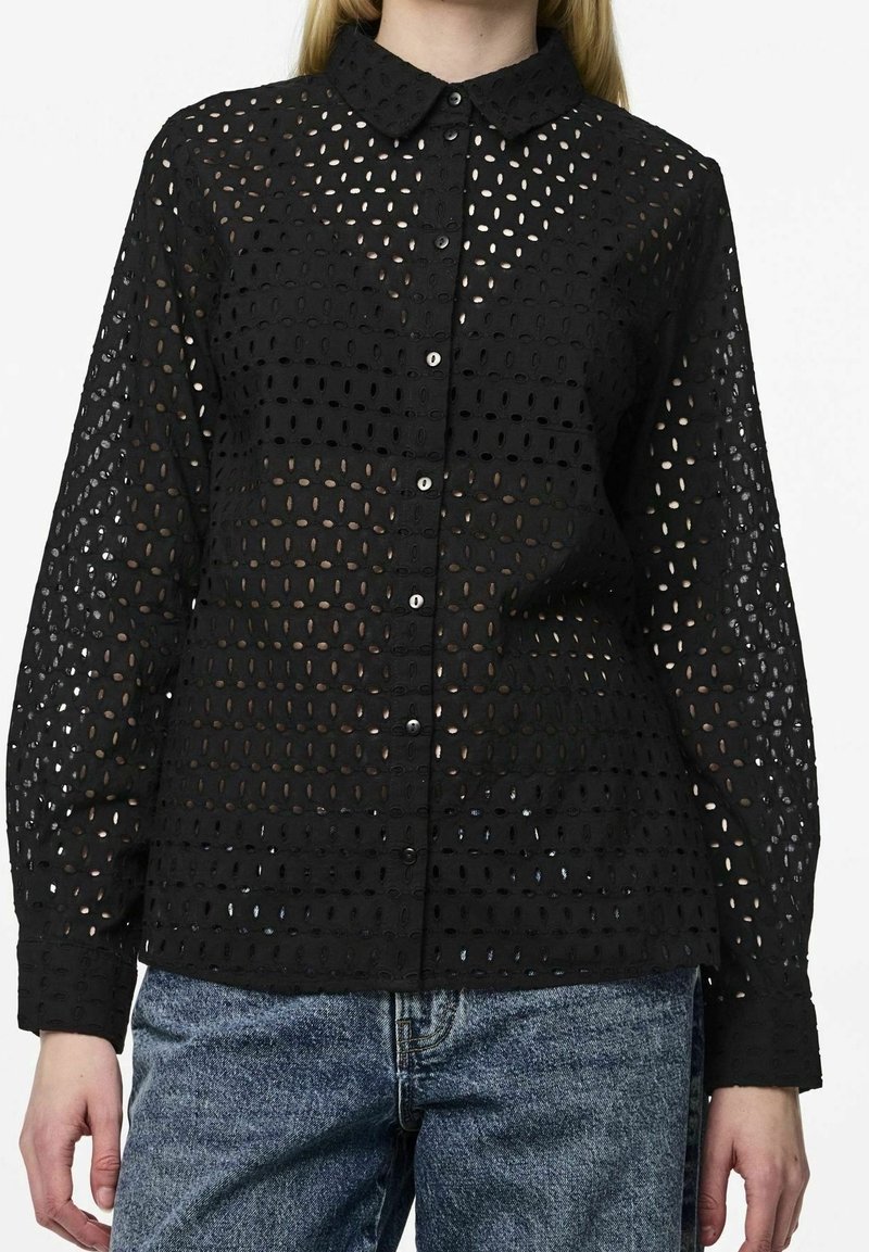 Černá košile na knoflíky vyrobená z vzorované krajkové tkaniny s oválnými výřezy. Má klasický límec a dlouhé rukávy, doplněná džínami.