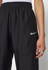Zwarte sportieve broek met een elastische tailleband, zijzakken en een zilveren Nike-logo op het linkerbeen. Gladde, lichte stof.