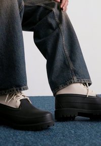 Zapatos de plataforma en negro y crema, de diseño grueso y con cordones, que cuentan con una suela de goma texturizada y un upper de cuero suave, sobre unos pantalones de mezclilla.