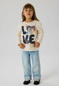 Kremfarget langermet skjorte med "LOVE" trykk som viser to tegneseriehunder, kombinert med lyseblå jeans og svarte slip-on sko.
