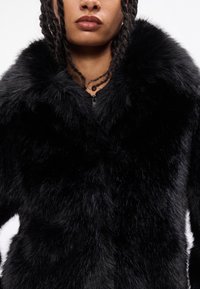 Casaco de pêlo sintético preto com colarinho alto, textura suave e fecho de zíper. O pêlo parece grosso e brilhante, com um design volumoso.