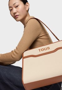 Bolso tote de lona con detalles en cuero marrón y asa superior. El bolso presenta un diseño curvo con el logo "TOUS" en relieve en la parte frontal.