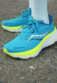 Chaussure de sport bleue avec une tige en maille texturée, des accents jaunes néon et une épaisse semelle blanche. Comprend un logo argenté et un design à lacets.