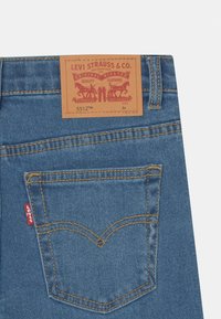 Denim jeans with a light blue wash, featuring a brown leather brand patch, yellow stitching, and a red logo tag on the back pocket.