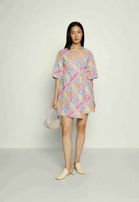 Mulher vestindo um vestido envolvente com padrão de patchwork em tons pastéis, segurando uma bolsa bege, combinada com sapatos de salto baixos bege, de pé sobre um fundo simples.