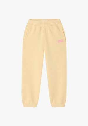 Dijete stoji, nosi preveliku narančastu majicu s natpisom "American Vintage", krem hlače i šarene sandale na jednobojnoj pozadini.