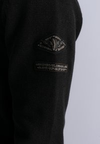 Black sweater fabric with textured knit. Left sleeve features a patch with an emblem and text label, featuring a dark color scheme.