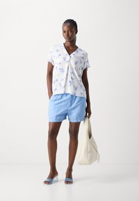Blusa floreale bianca con motivi blu, pantaloni corti azzurri e tacchi aperti blu. Borsa a spalla beige. Tessuto morbido, vestibilità rilassata.
