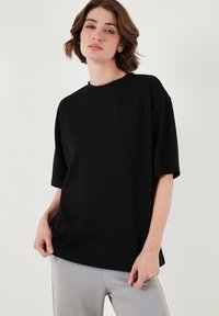 Melna oversized t-krekls, kas izgatavots no mīkstas auduma, ar apaļu kakla izgriezumu un īsām piedurknēm, apvienots ar gaiši pelēkiem biksēm. Vienkāršs un minimālistisks dizains.