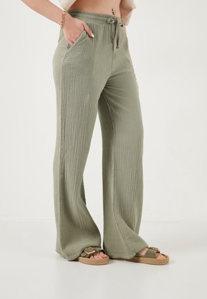 Pantaloni ampi in verde oliva con trama e vita con coulisse, indossati con sandali abbinati e braccialetto su sfondo chiaro.
