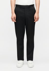 Άνδρας με στενά μαύρα παντελόνια, λευκά αθλητικά παπούτσια και λευκό πουκάμισο που είναι φορεμένο μέσα, στέκεται μπροστά σε απλό ανοιχτόχρωμο φόντο.