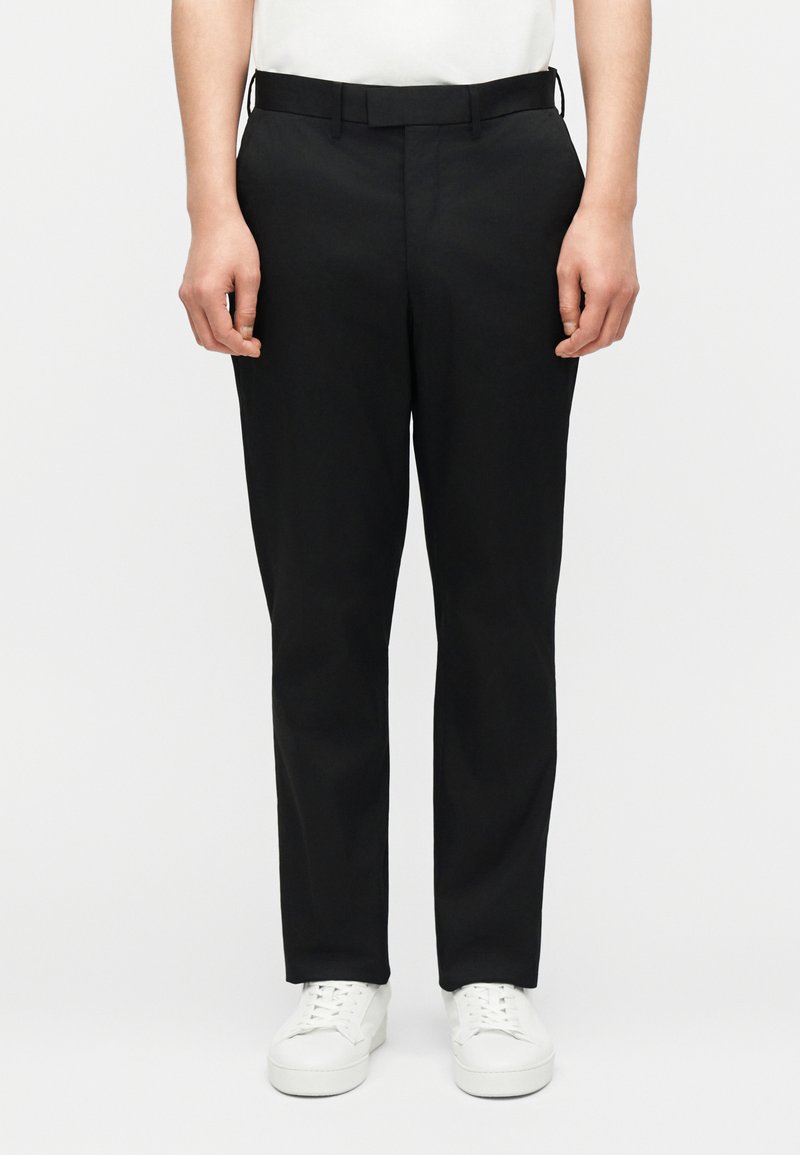 Άνδρας με στενά μαύρα παντελόνια, λευκά αθλητικά παπούτσια και λευκό πουκάμισο που είναι φορεμένο μέσα, στέκεται μπροστά σε απλό ανοιχτόχρωμο φόντο.