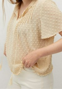 Light beige blouse with textured polka dot pattern, flutter sleeves, front tassel ties, and scalloped hem worn with white pants.