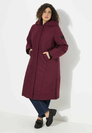 Bordeaux lange pufferjas met een capuchon, ritssluitingen aan de zijkanten en zwarte accenten. Gecombineerd met donkere jeans en zwarte chunky schoenen.